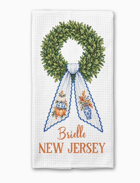 Personalized chinoiserie fall kitchen towel featuring a green boxwood wreath, blue and white ribbon, pumpkins and custom Brielle New Jersey text.