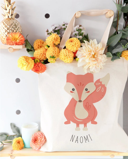 White tote bag with a fox design and 'Naomi' text, surrounded by flowers and decorative items.