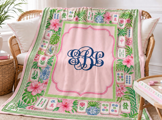 Personalized pink Mahjong minky fleece blanket with navy interlocking monogram, tropical flowers, green leaves and colorful Mahjong tile border displayed beside a Mahjong table.