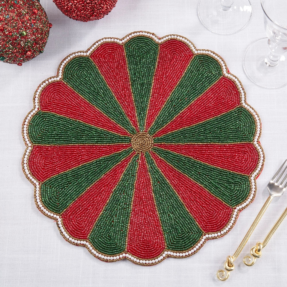 A round scalloped beaded placemat with alternating red and green segments and a gold-toned center, styled on a white tablecloth with gold-toned flatware and holiday decorations.