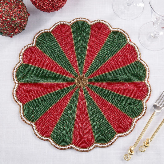 A round scalloped beaded placemat with alternating red and green segments and a gold-toned center, styled on a white tablecloth with gold-toned flatware and holiday decorations.
