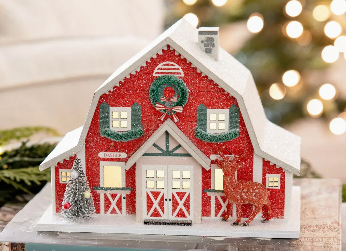 Decorative Christmas barn with lights, wreath, and deer in front of a blurred festive background.