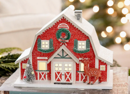 Decorative Christmas barn with lights, wreath, and deer in front of a blurred festive background.