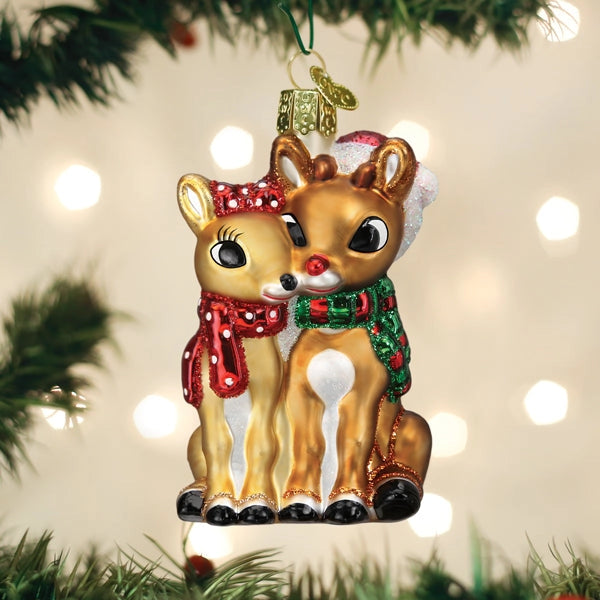 A Christmas ornament shaped like two kissing reindeer hanging from a green tree branch. One reindeer wears a red scarf with white polka dots and a Santa hat, while the other wears a green patterned scarf.