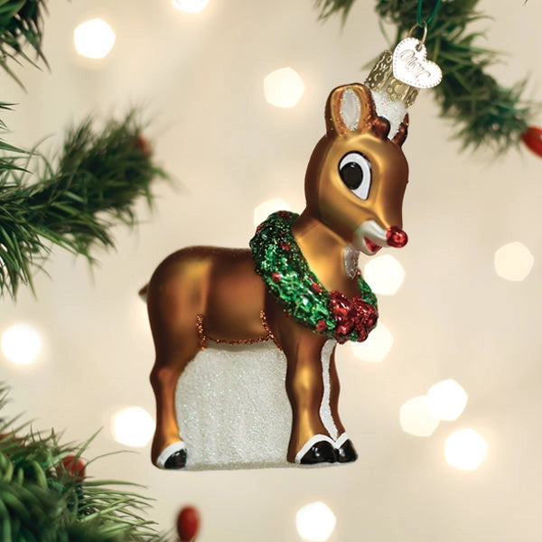 A glossy brown Christmas ornament shaped like a reindeer, decorated with a green and red glittery wreath, a white hooves patch, and a silver hanging loop, shown hanging against a soft-focus background with pine branches and warm bokeh lights.