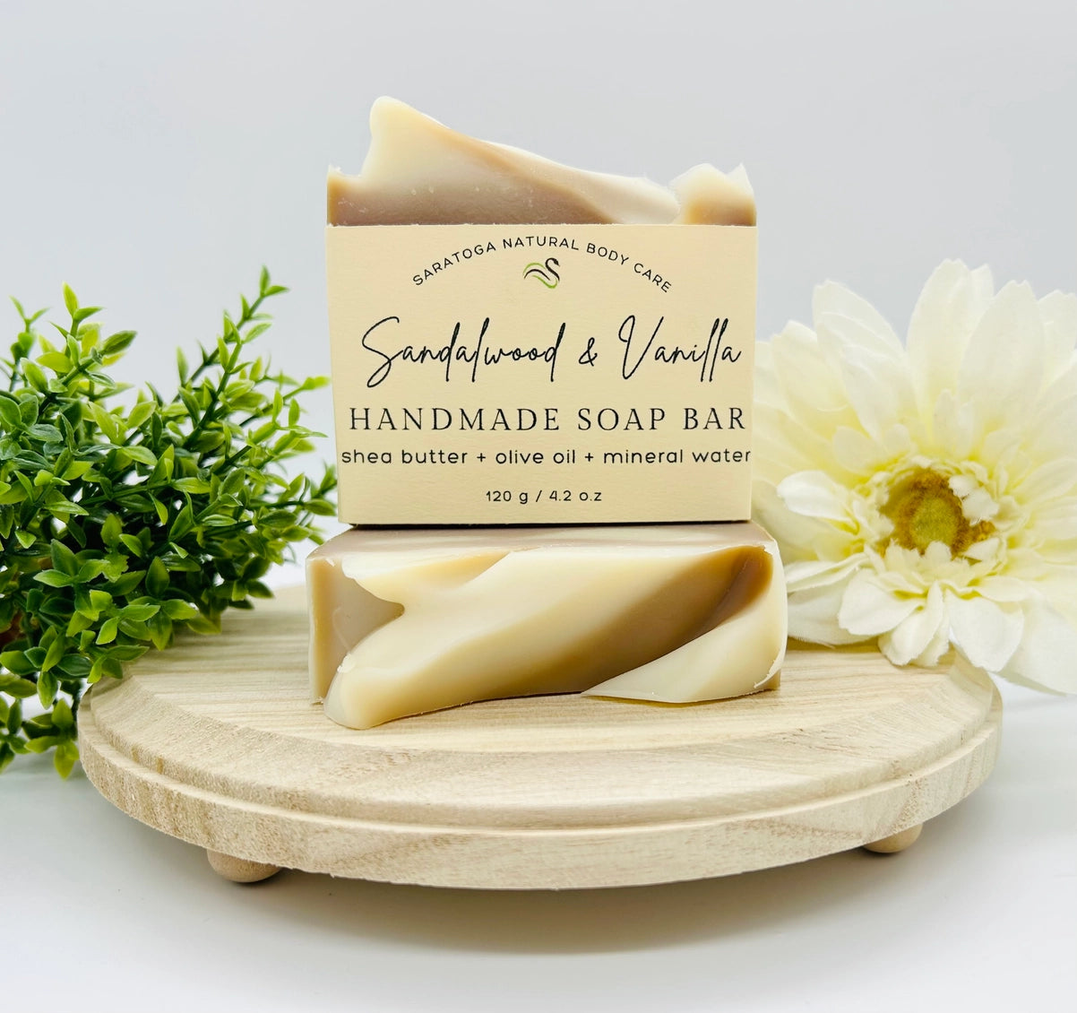 Handmade soap bar with packaging on a wooden stand, surrounded by flowers and greenery.