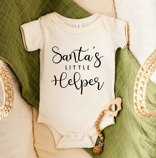 Baby onesie with 'Santa's Little Helper' text on a green blanket with a teething ring.