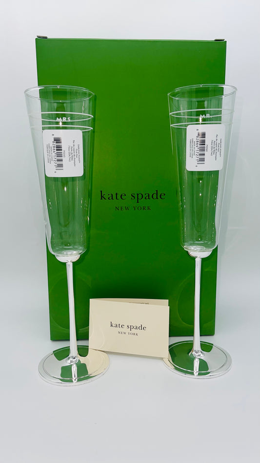 Two champagne flutes with a green box and card featuring 'kate spade' branding on a white background.
