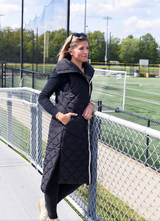 Woman wearing a long black quilted coat standing on a tennis court.