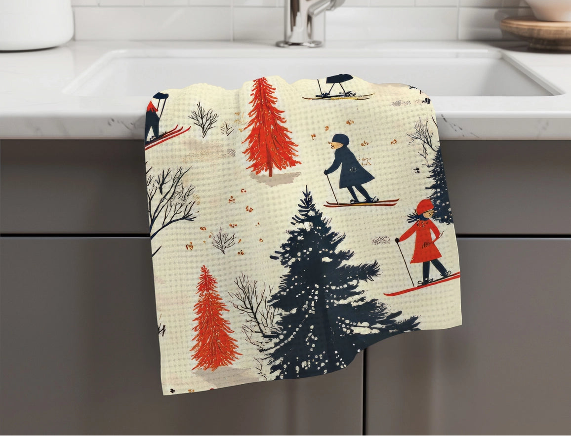A kitchen towel with a waffle texture hangs over the edge of a white sink. The towel features a whimsical winter print of skiers, pine trees, and bare winter trees in navy, red, and beige.