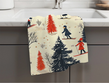 A kitchen towel with a waffle texture hangs over the edge of a white sink. The towel features a whimsical winter print of skiers, pine trees, and bare winter trees in navy, red, and beige.