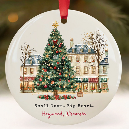 Round white ceramic Christmas ornament featuring a winter town scene with a decorated Christmas tree, historic buildings, bare trees, and the text 'Small Town. Big Heart.