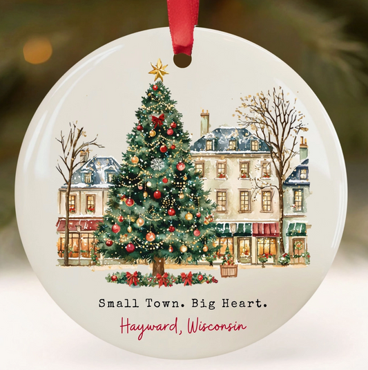 Round white ceramic Christmas ornament featuring a winter town scene with a decorated Christmas tree, historic buildings, bare trees, and the text 'Small Town. Big Heart.