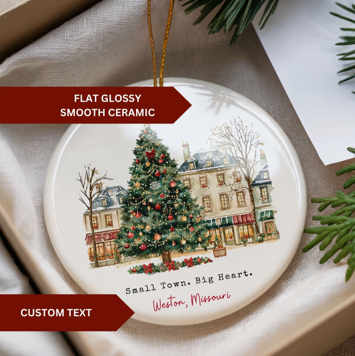 A round glossy ceramic Christmas ornament displayed in a gift box, featuring an illustrated winter street scene with a decorated Christmas tree and the text 'Small Town. Big Heart.
