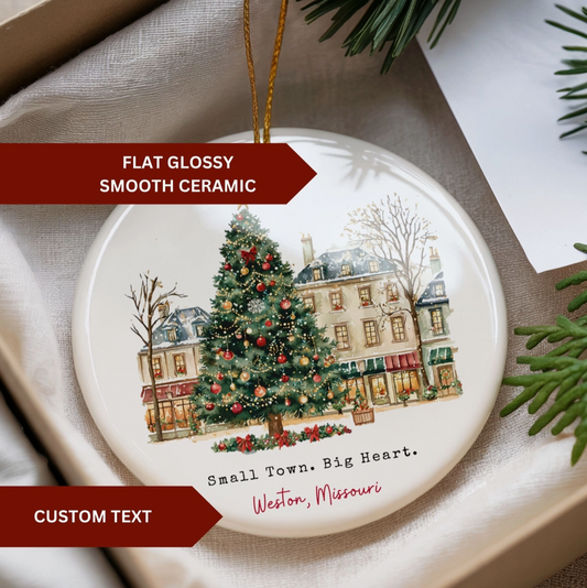 A round glossy ceramic Christmas ornament displayed in a gift box, featuring an illustrated winter street scene with a decorated Christmas tree and the text 'Small Town. Big Heart.