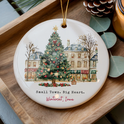 A round ceramic Christmas ornament with a gold hanging loop, featuring an illustrated holiday street scene with a decorated tree, buildings, and the text 'Small Town. Big Heart.