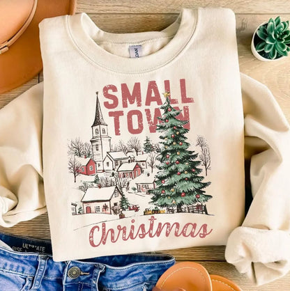 Beige sweatshirt with 'Small Town Christmas' design featuring a village and Christmas tree on a wooden surface.