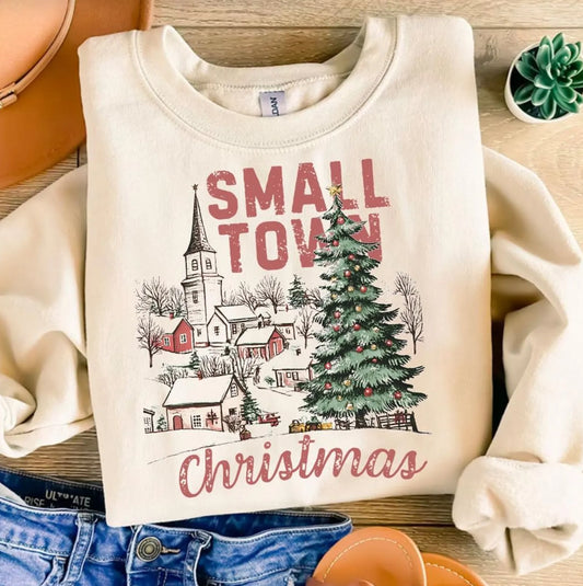 Beige sweatshirt with 'Small Town Christmas' design featuring a village and Christmas tree on a wooden surface.