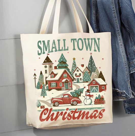 A cream canvas tote bag with a festive Christmas winter village print, featuring the words 'SMALL TOWN Christmas' in green and red lettering. The bag is hanging next to a pair of blue denim jeans against a gray paneled wall.