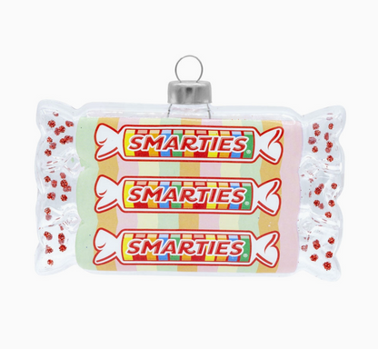 A novelty Christmas ornament shaped like a Smarties candy bar wrapper, featuring three printed Smarties logos and colorful striped details, with a silver hanging loop at the top.