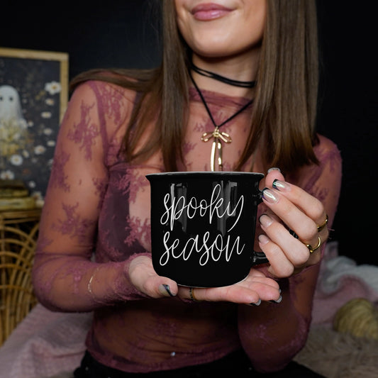 Person holding a black mug with 'Spooky Season' text in a dark setting