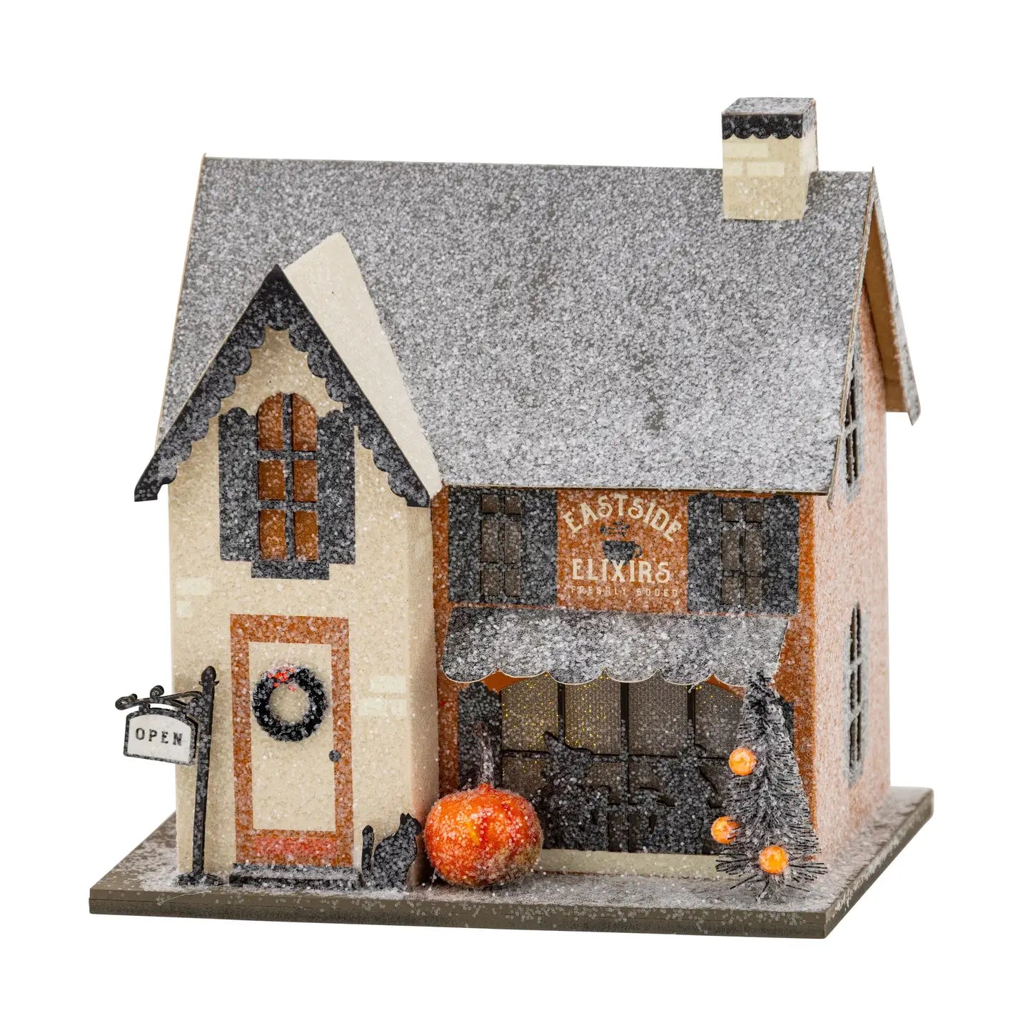 A decorative miniature house with a snowy gray roof, beige and orange facade, a storefront sign reading "Eastside Elixirs," a small pumpkin, a black cat figurine, and an open sign on a white background.