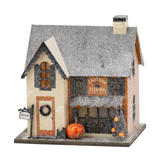 A decorative miniature house with a snowy gray roof, beige and orange facade, a storefront sign reading "Eastside Elixirs," a small pumpkin, a black cat figurine, and an open sign on a white background.