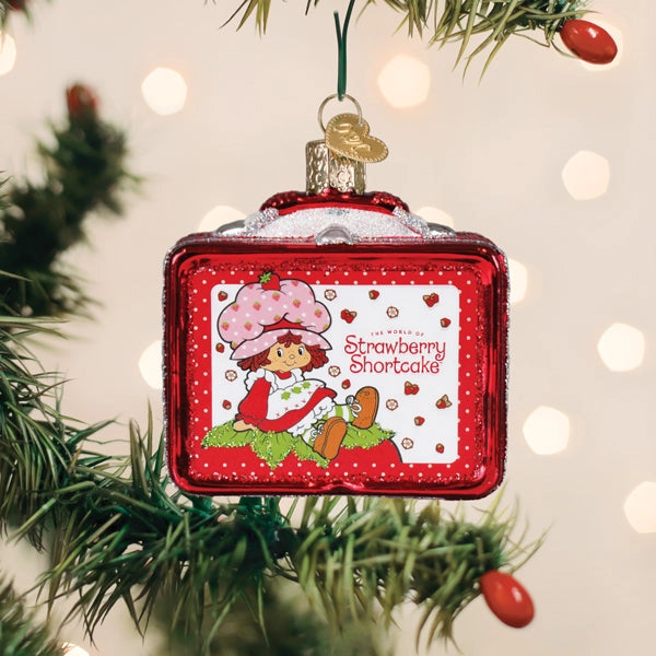 A Strawberry Shortcake Christmas ornament shaped like a red suitcase, hanging from a green Christmas tree branch with warm bokeh lights in the background.
