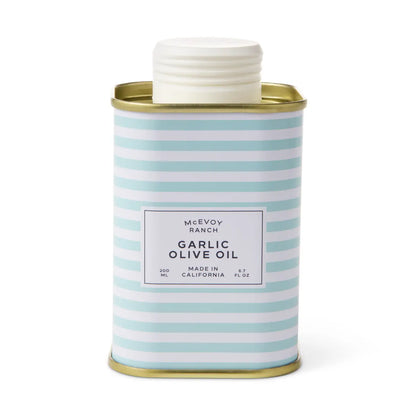 A McEvoy Ranch Garlic Olive Oil canister with a white screw cap, featuring a light blue and white striped body with a white label and gold metal trim, set against a plain white background.