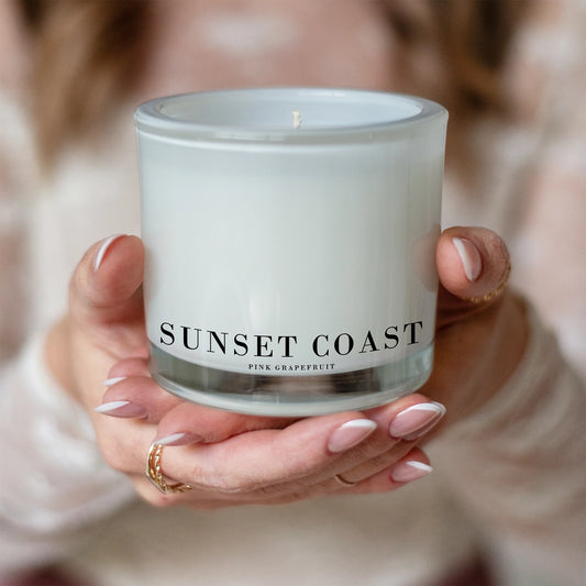 Hand holding a candle labeled 'Sunset Coast' with a soft focus background