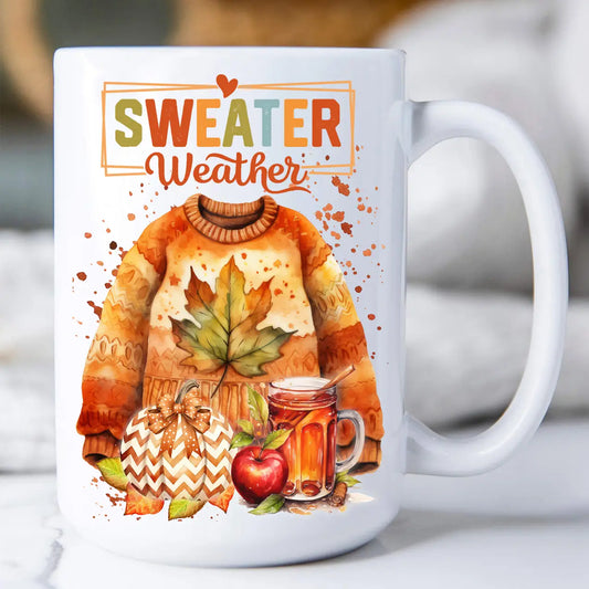 A white ceramic mug featuring a colorful autumn-themed graphic with the text 'SWEATER Weather' above an illustrated orange sweater with a maple leaf, a pumpkin, an apple, and a mug of iced tea, set against a softly blurred indoor background.