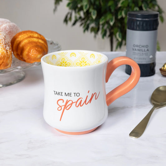 White mug with orange handle and 'Take Me to Spain' text on a table with coffee and pastries.