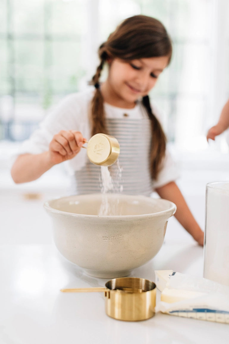 Child pouring liquid from a measuring cup into a mixing bowl on a white surface.