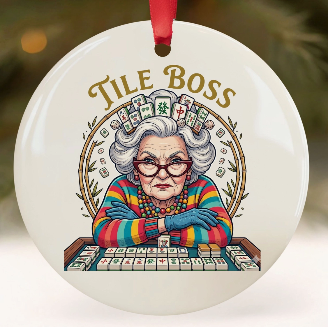 A round white ceramic Christmas ornament featuring a colorful illustration of an elderly woman at a mahjong table with the words 'TILE BOSS' above her. The ornament has a red hanging ribbon and is shown against a softly blurred festive background.