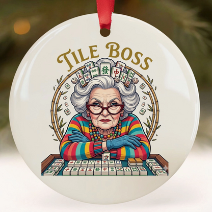 A round white ceramic Christmas ornament featuring a colorful illustration of an elderly woman at a mahjong table with the words 'TILE BOSS' above her. The ornament has a red hanging ribbon and is shown against a softly blurred festive background.