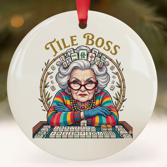 A round white ceramic Christmas ornament featuring a colorful illustration of an elderly woman at a mahjong table with the words 'TILE BOSS' above her. The ornament has a red hanging ribbon and is shown against a softly blurred festive background.