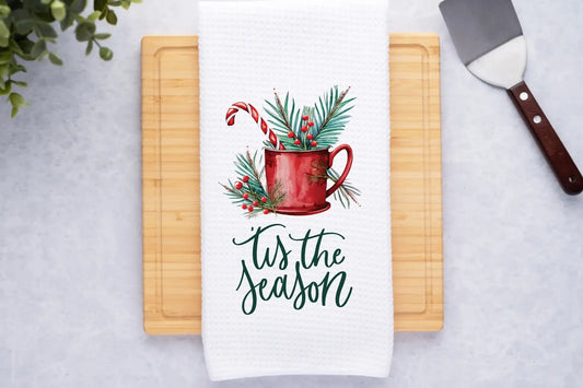 Towel with Christmas design and 'tis the season text on a cutting board.