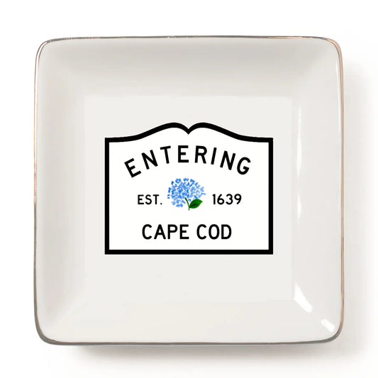 A square white ceramic dish with rounded corners and a metallic gold-toned rim, featuring a printed black-bordered label in the center that reads 'ENTERING CAPE COD EST. 1639' with a small blue flower graphic.