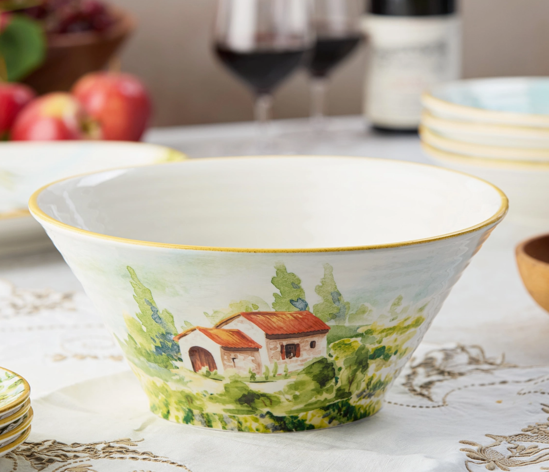 Decorative ceramic bowl with landscape design on a table with wine glasses and apples in the background