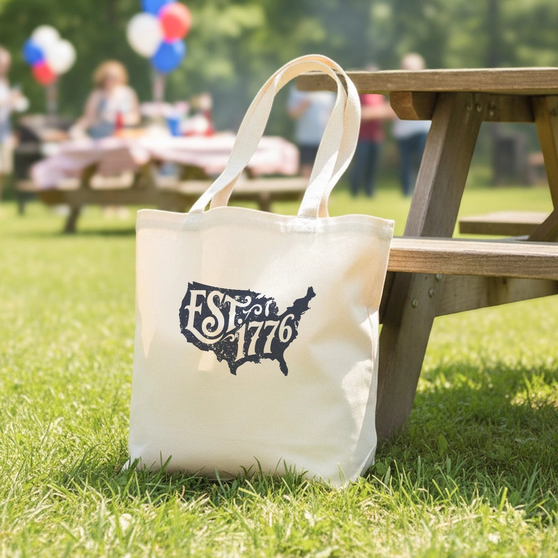 White tote bag with 'EST 1776' design on grass next to a wooden picnic table