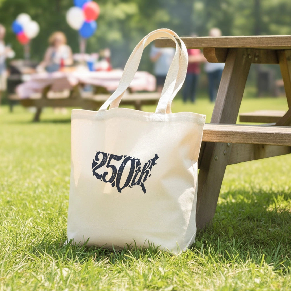 White tote bag with '250th' text on grass next to a wooden picnic table with blurred people and balloons in the background.