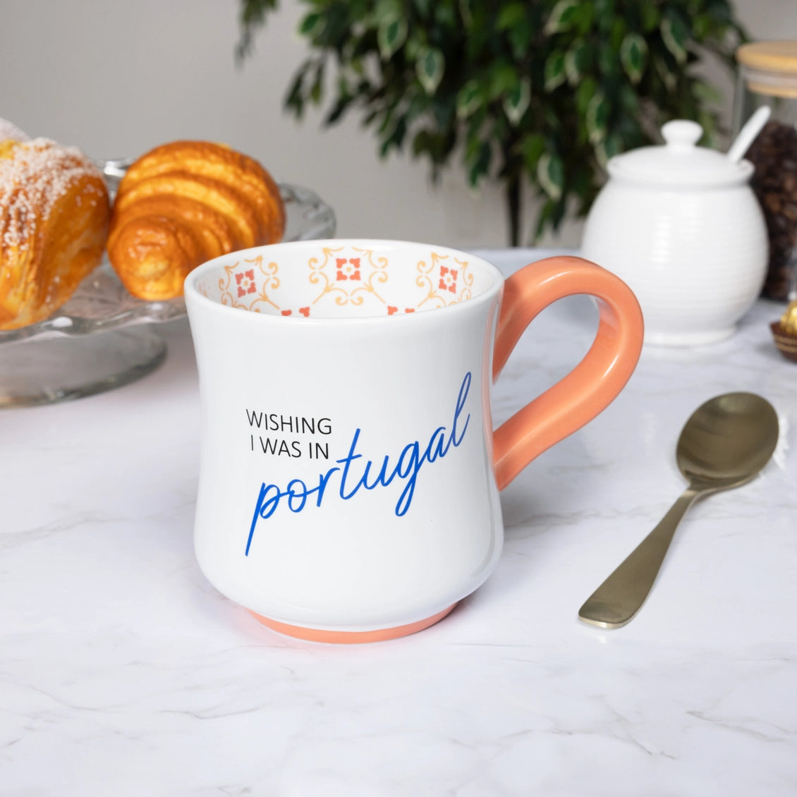 Mug with 'Wishing I was in Portugal' text on a table with pastries and a teapot.