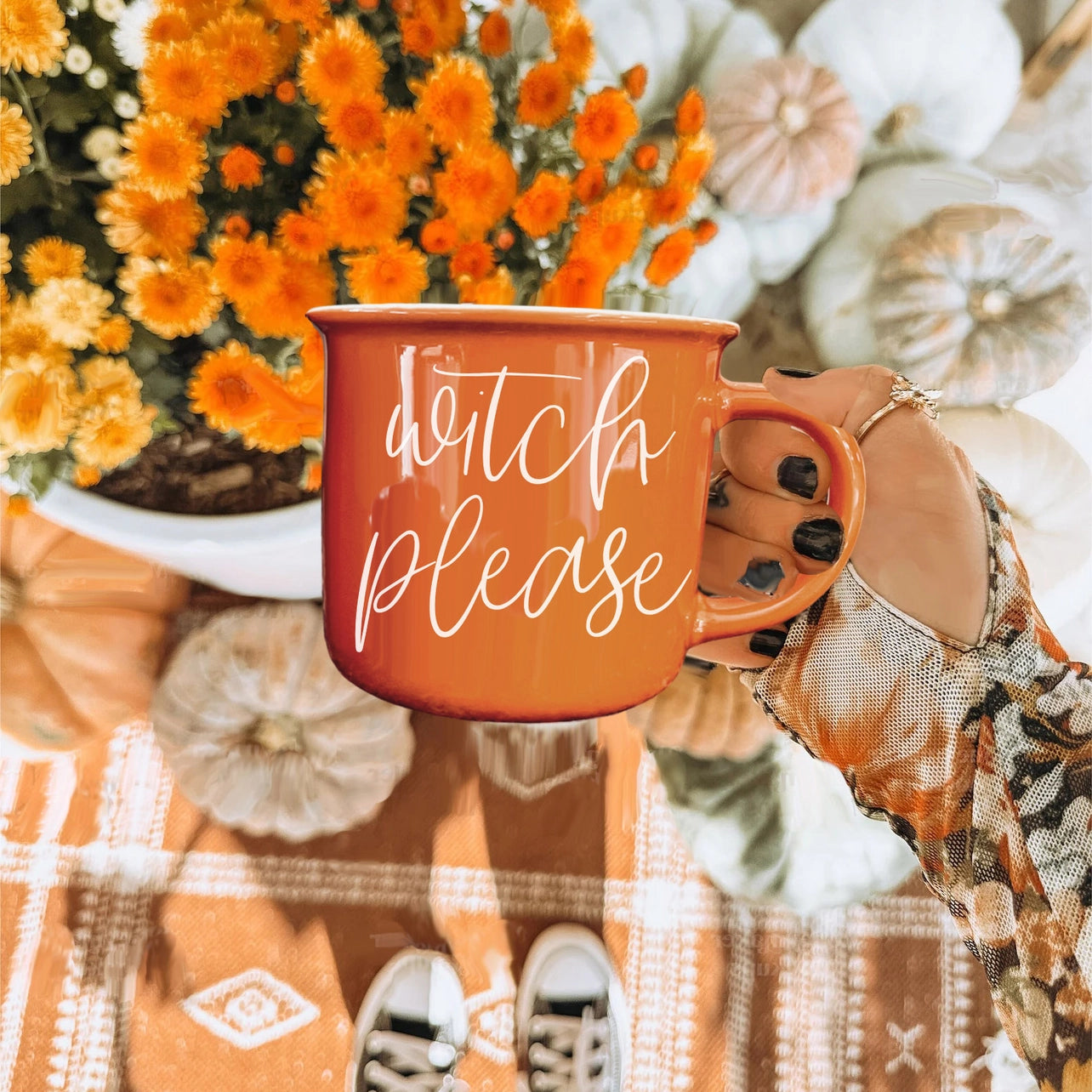Orange mug with 'witch please' text held by a hand, surrounded by autumn decorations.