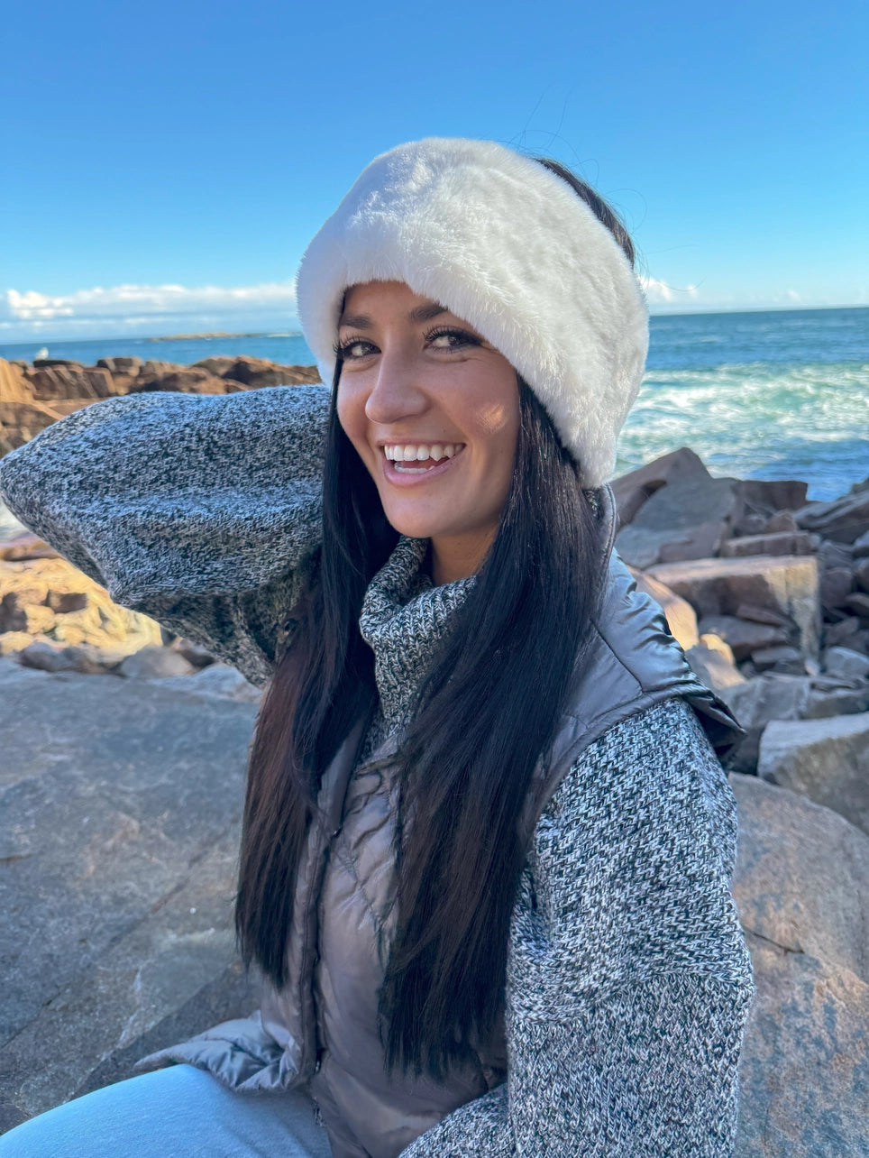 Woman wearing a white Faux Fur headband and gray vest sitting on rocks by the ocean.