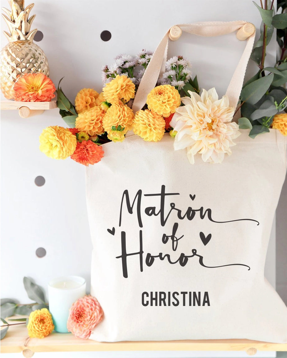 White tote bag with 'Matron of Honor Christina' text, surrounded by flowers and decor.