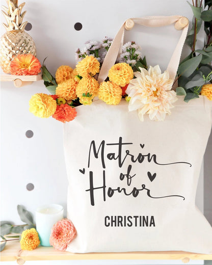 White tote bag with 'Matron of Honor Christina' text, surrounded by flowers and decor.