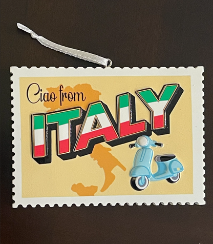 A square decorative ornament or plaque with a white scalloped border and hanging ribbon, featuring the words 'Ciao from ITALY' in large letters filled with the Italian flag colors, a light blue scooter, and an orange map silhouette of Italy on a pale yellow background, photographed on a dark brown surface.