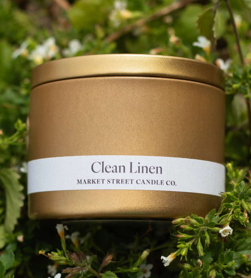 Market Street Candle Co. Clean Linen Travel Tin soy candle in a gold metal tin surrounded by green foliage and small white flowers.

