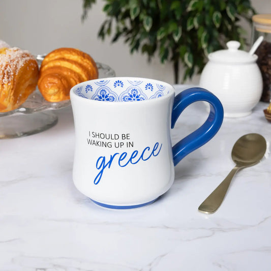 White mug with blue handle and text 'I should be waking up in Greece' on a table with pastries and a teapot.